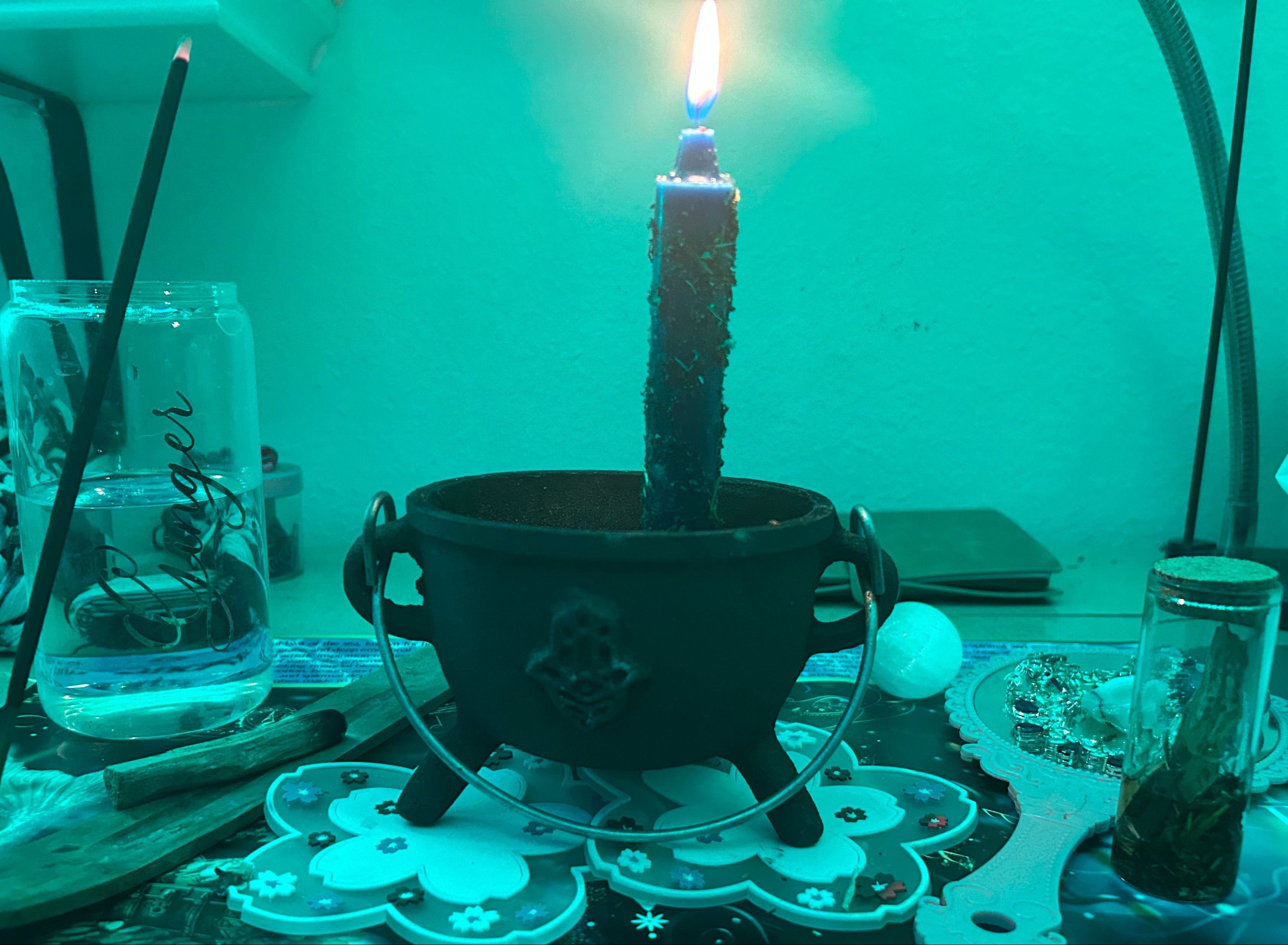 Candle in a cauldron with mystical paraphernalia on a table, illuminated by a green light.